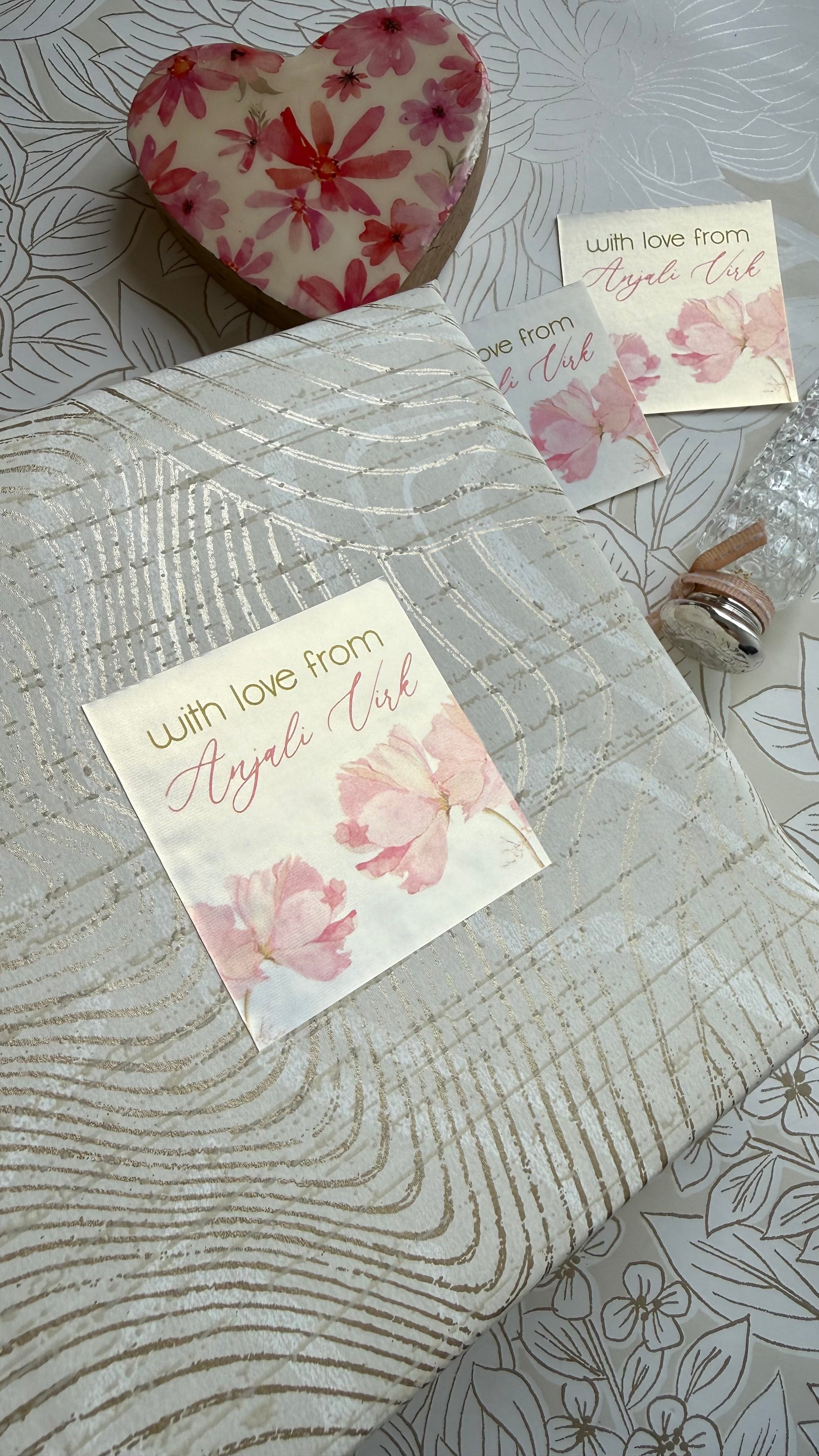 A gift-wrapped package with a floral tag reading 'with love from Angel's Kid' placed on a patterned surface, alongside a heart-shaped floral item and small cards.