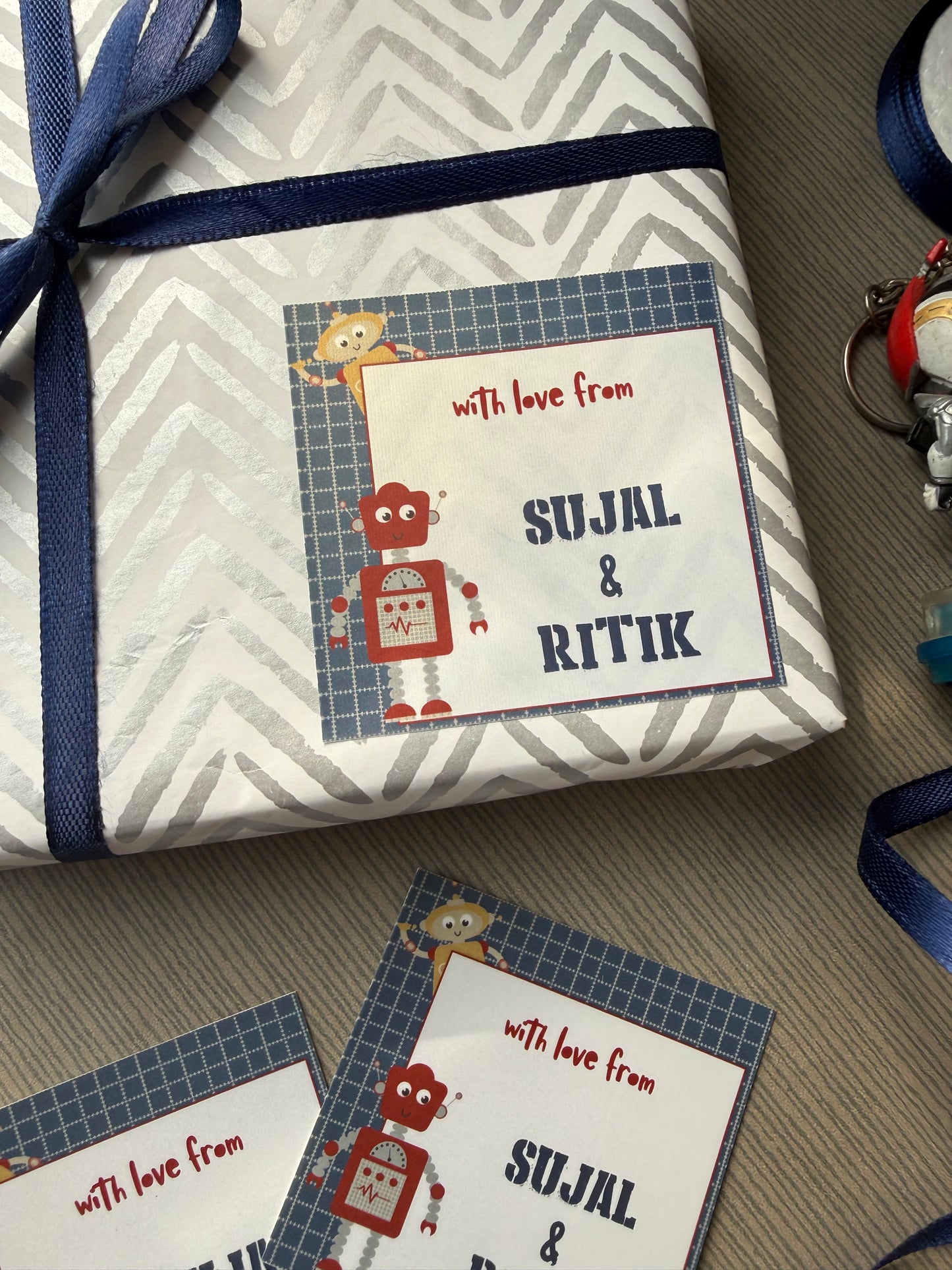 A wrapped gift box with a personalized card that reads 'with love from SUJAL & RITIK' and 'SULJAL & RITIK' in large letters, tied with a navy ribbon, on a gray carpet. Another matching card is placed beside it.