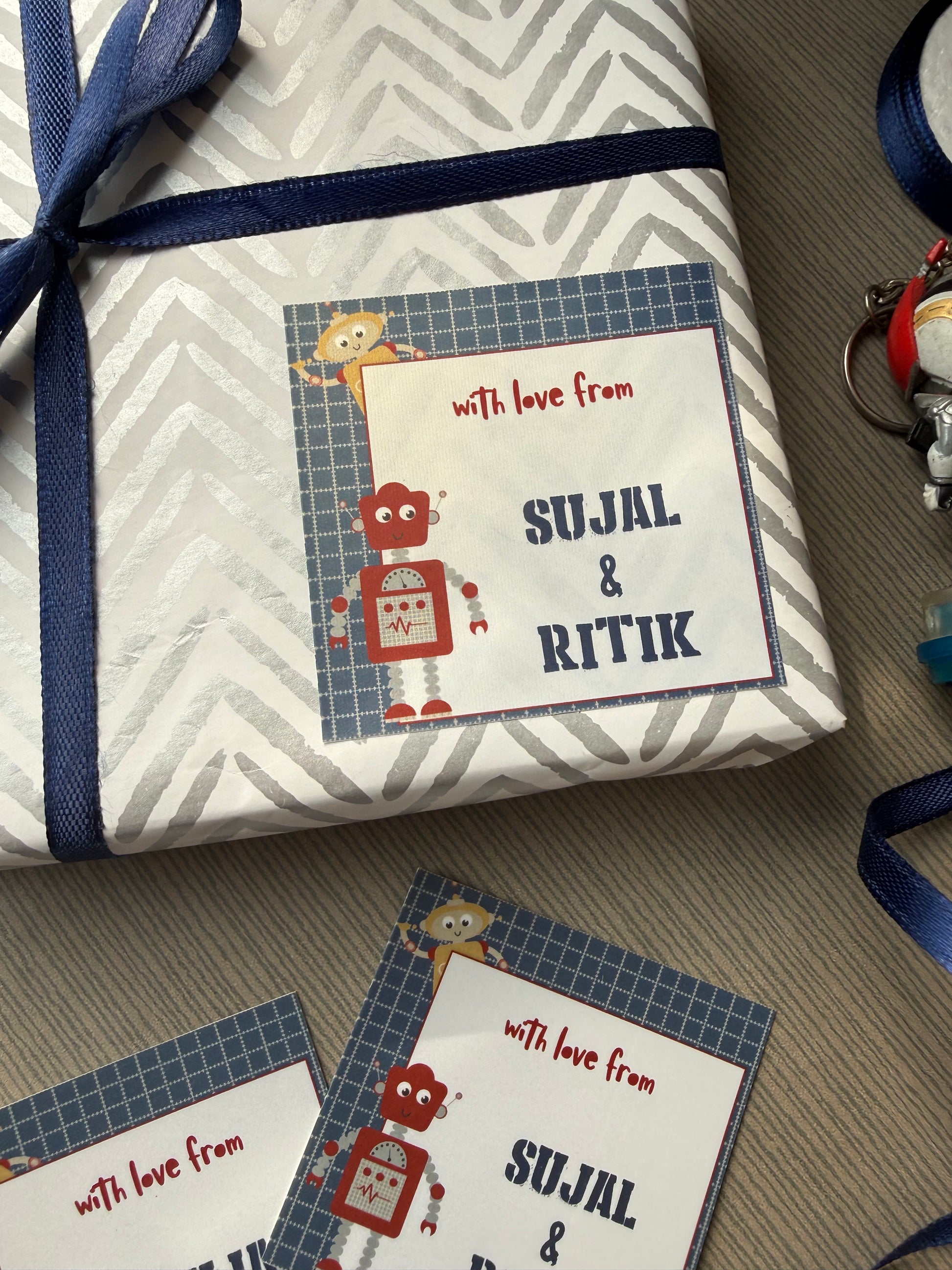 A wrapped gift box with a personalized card that reads 'with love from SUJAL & RITIK' and 'SULJAL & RITIK' in large letters, tied with a navy ribbon, on a gray carpet. Another matching card is placed beside it.