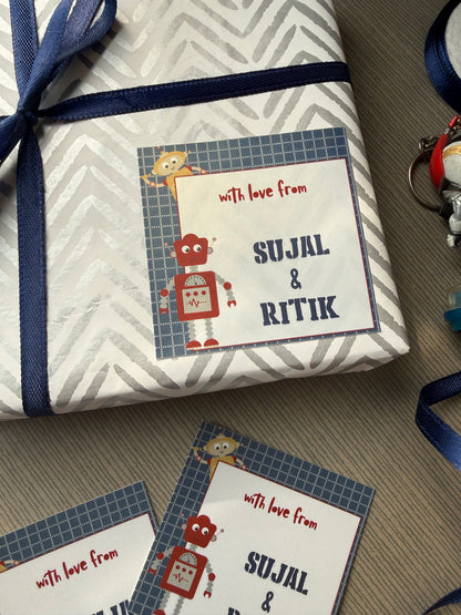 A wrapped gift box with a personalized card that reads 'with love from SUJAL & RITIK' and 'SULJAL & RITIK' in large letters, tied with a navy ribbon, on a gray carpet. Another matching card is placed beside it.
