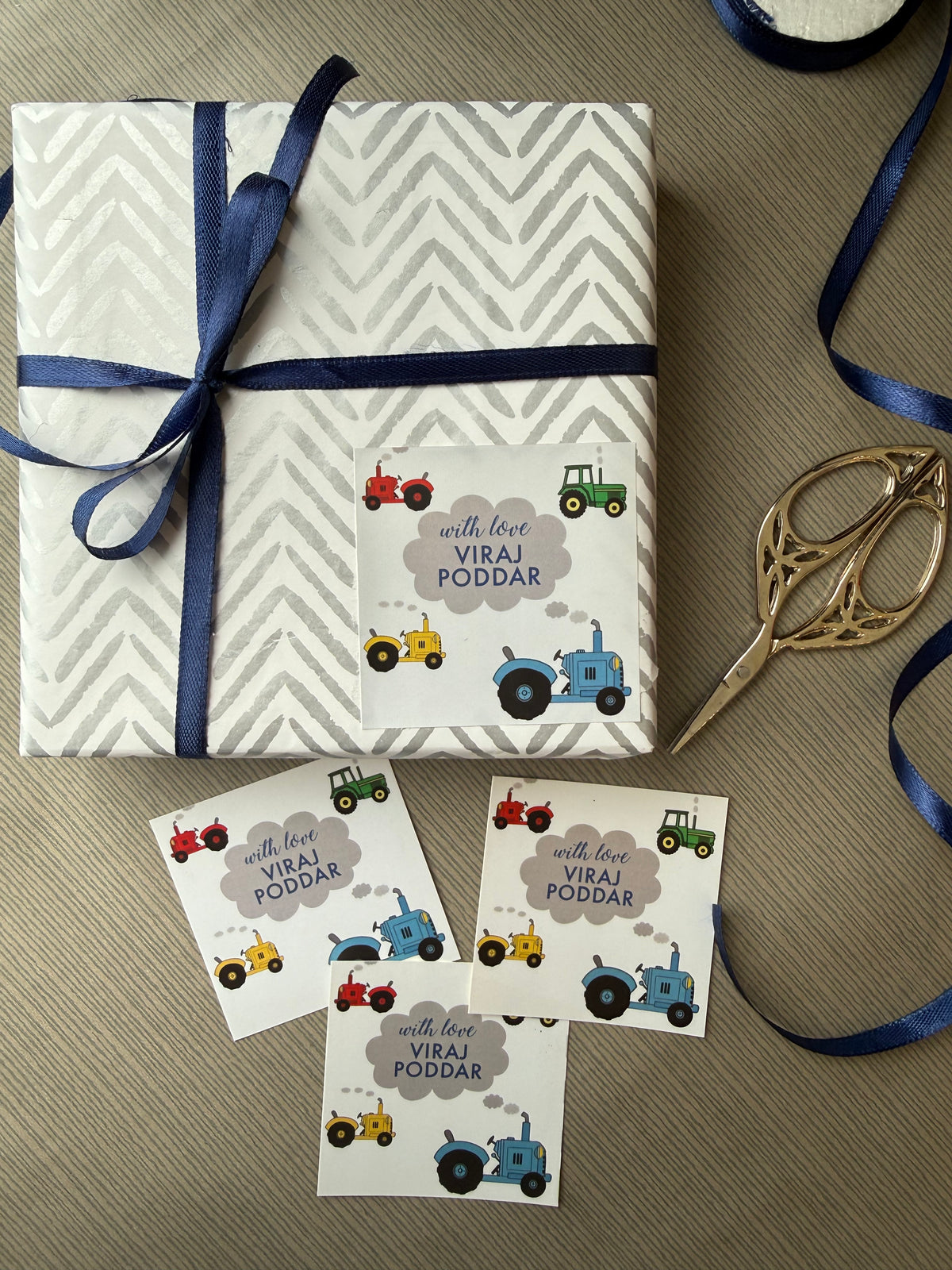 A wrapped gift box and matching printed cards with tractor illustrations, tied with a navy ribbon, arranged on a textured beige surface with scissors and ribbon visible around it.