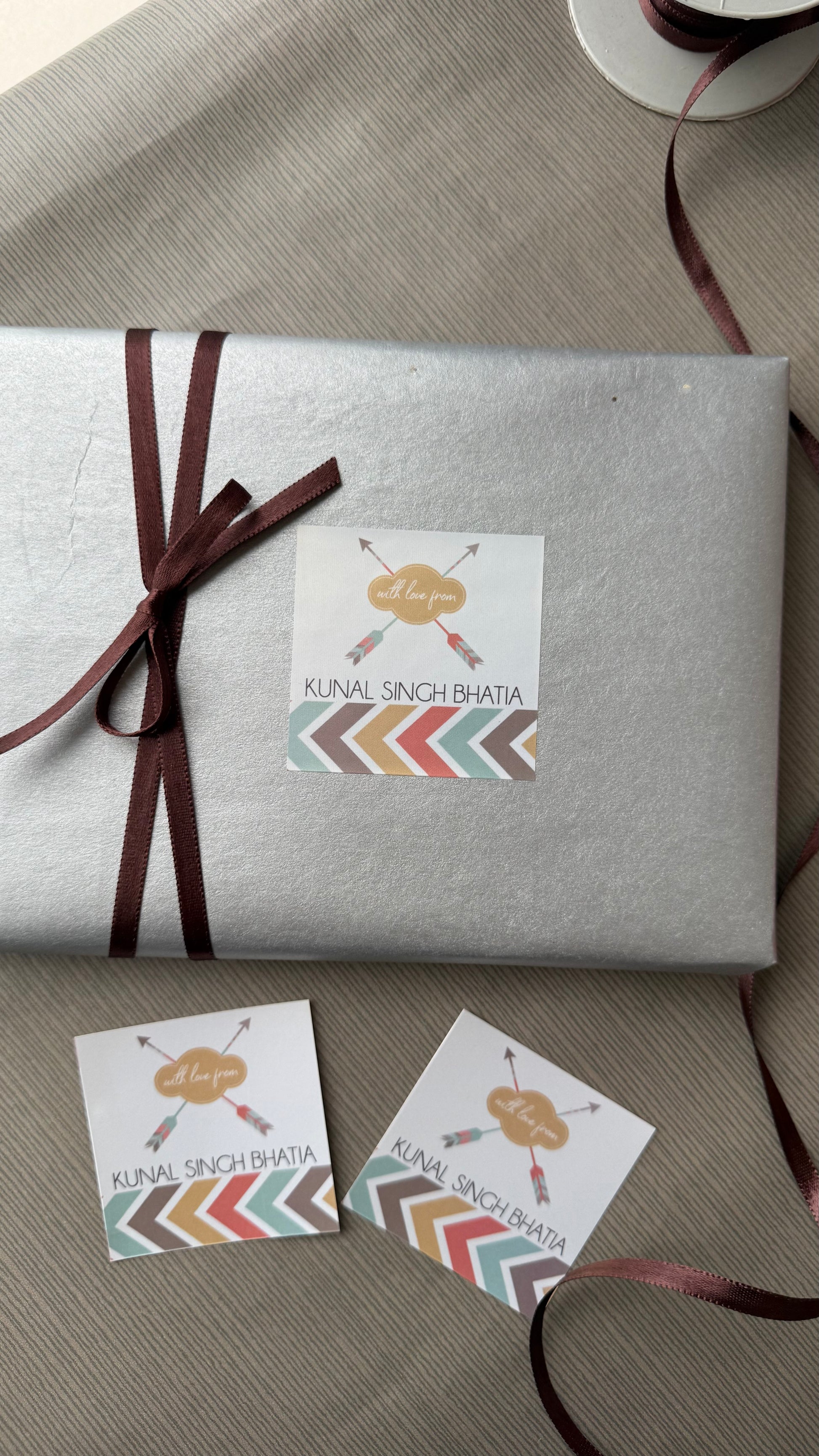 A silver gift-wrapped box tied with a dark brown ribbon, featuring a square printed label that reads 'KUNAL SINGH BHAIYA' with crossed arrows and a multicolor chevron design. Two matching labels are placed on the table beside it.