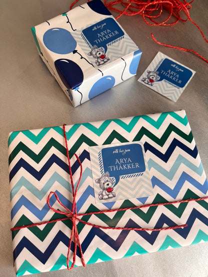 Personalized gift wrap and matching tags featuring a boy's birthday theme with the name 'Arya Thakker' and a cartoon dog illustration. The main gift box in the foreground has a blue, green, and white chevron pattern and is tied with red string.