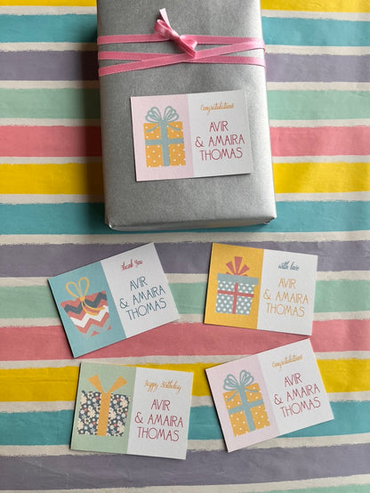 A wrapped gift box tied with a pink ribbon sits on a pastel striped surface, with four small gift tag cards arranged below. The tags have birthday greetings 