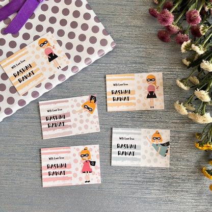 Four rectangular gift tags with cartoon superhero illustrations and the text 'Wish From RASHMI RAWAT' are arranged on a gray wood surface next to a polka-dot gift bag and dried flowers.