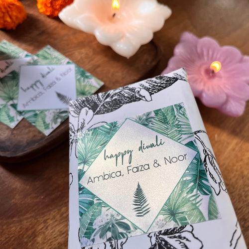 Gift labels with 'Happy Diwali' text and decorative elements in gold and silver colors, presented on a wooden board with candles and flower accents in the background.