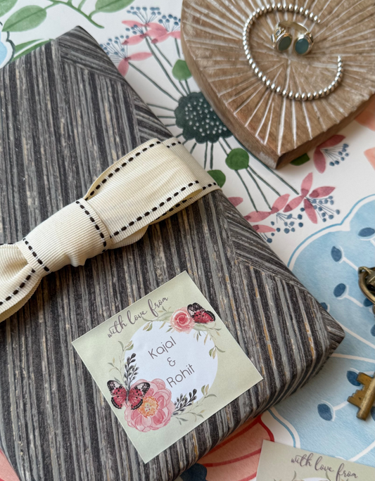 A wrapped gift box with a dark gray wood-grain patterned paper, tied with a cream ribbon, and adorned with a floral 'with love from Kaja & Rohi' sticker. T