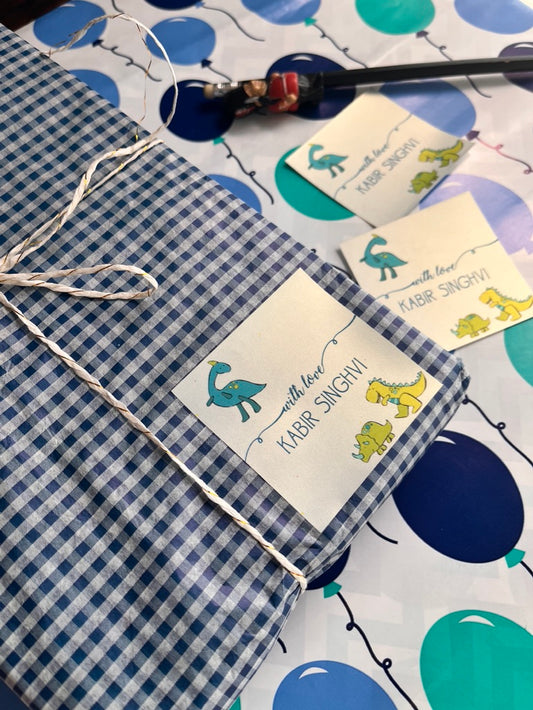 A folded blue-and-white gingham fabric item tied with white string, with a personalised gift label that reads 'with love KABIR SINGHAV' and small dinosaur illustrations. Other matching labels are visible on a balloon-patterned surface.