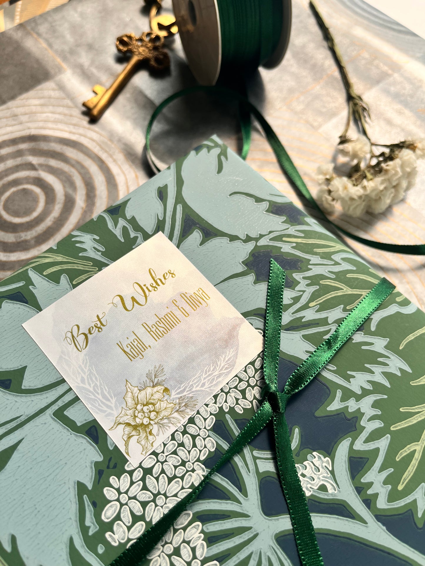 A close-up of a wrapped gift box with a botanical patterned paper sleeve in sage green, navy, and white, tied with a dark green ribbon and a small white tag with gold script.