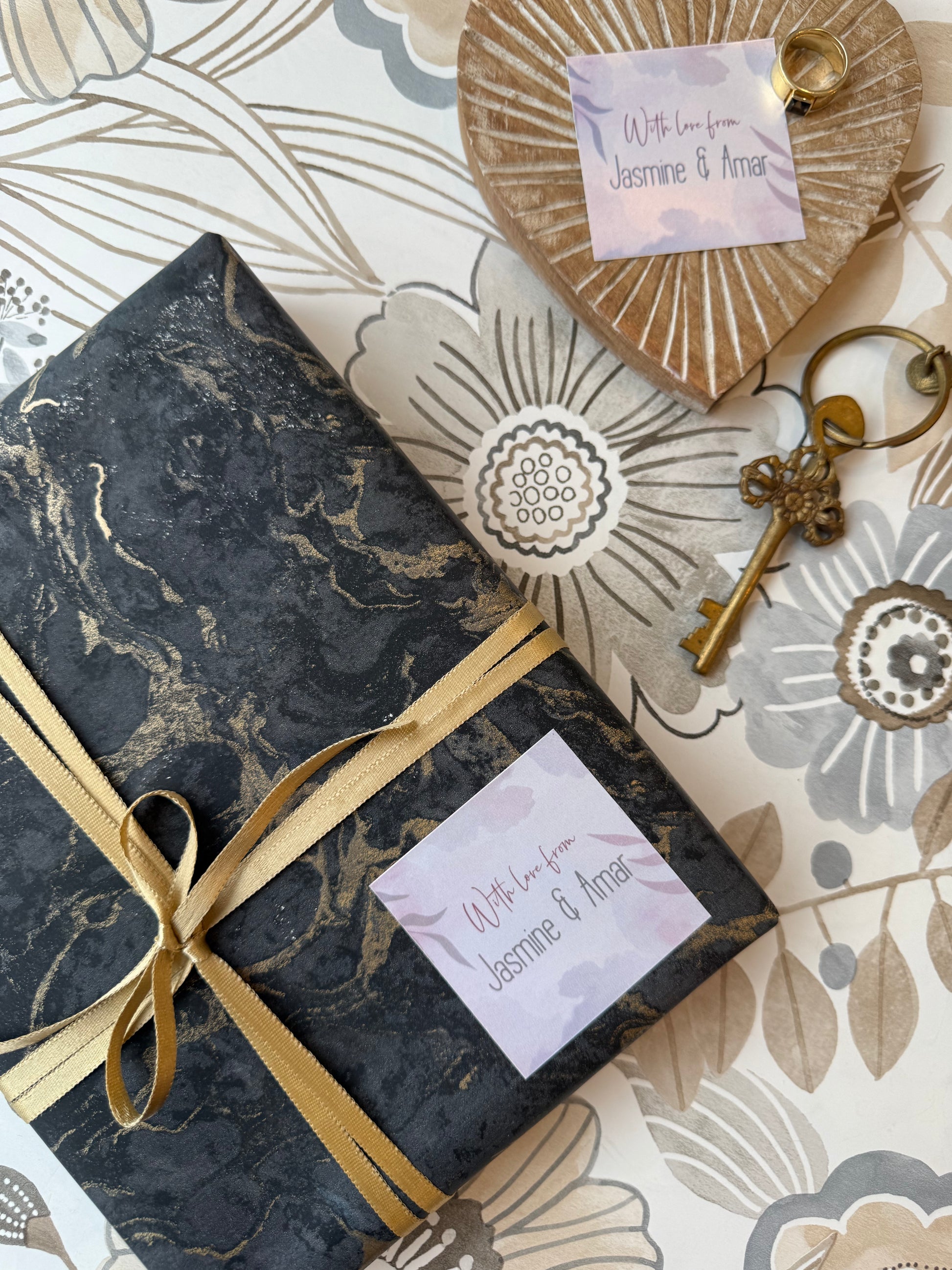 A wrapped gift box with a black and gold marble pattern, tied with a gold ribbon, to a wooden heart tag, a vintage-style key, and a personalised gift label reading 'With love from Jasmine & Amra'.