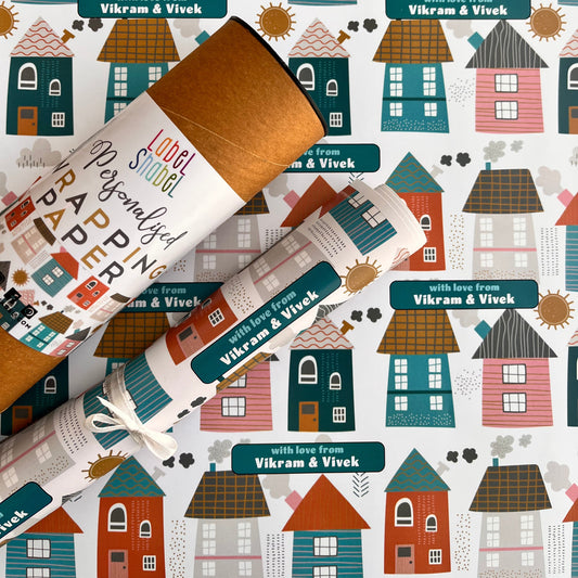 Personalized wrapping paper featuring a whimsical house pattern in teal, terracotta, brown, and cream on a white background, shown with two rolled tubes of kraft paper.