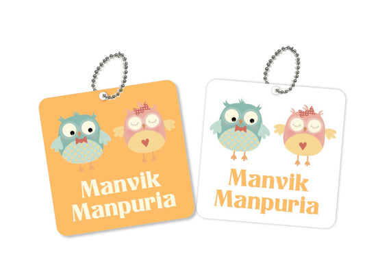 Two square metal bag tags with an owl design, one orange and one white, both with the name 'Manvik' printed on them.