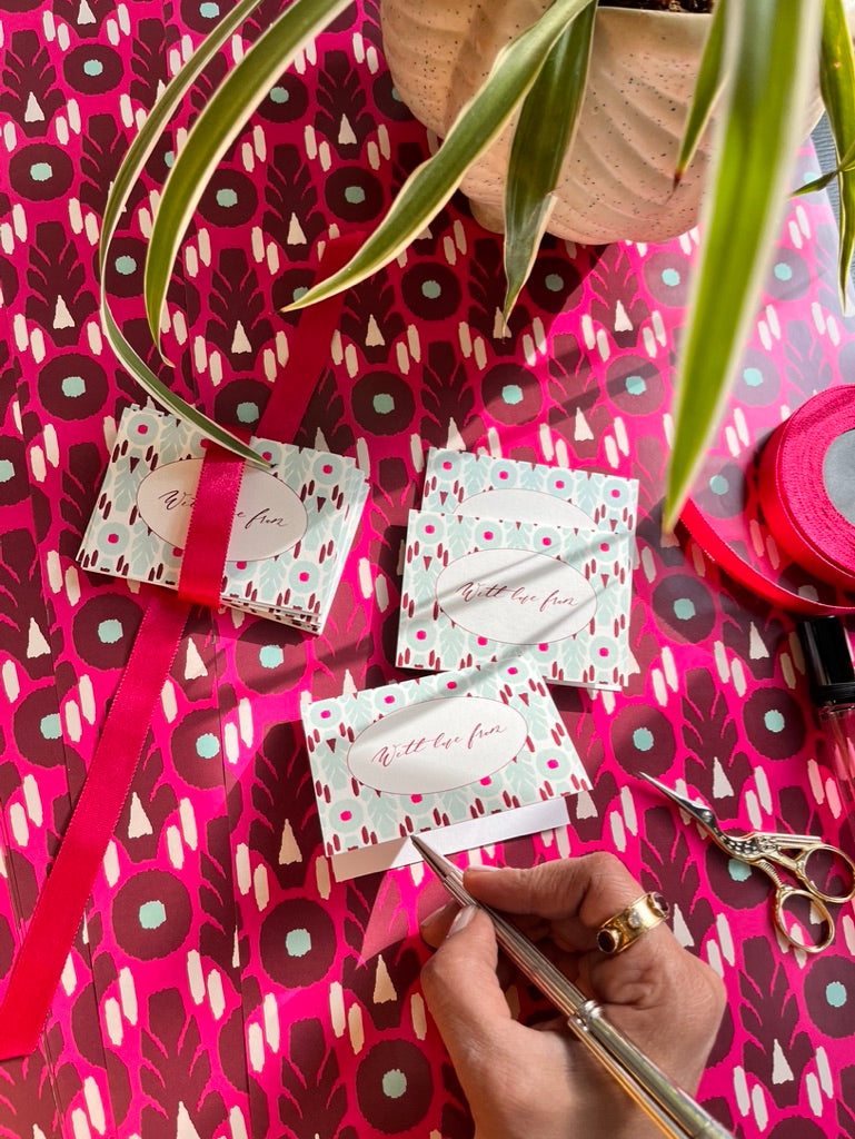 A CLOSE UP OF A HAND WRITING OUT A MESSAGE IN A GIFT TAG IN BRIGHT PINK IKAT DESIGN