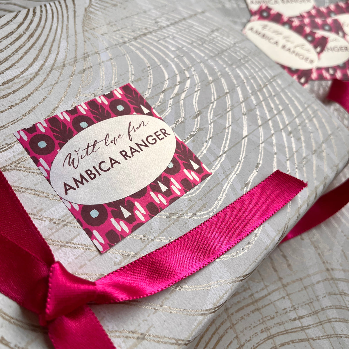 A close-up of a wrapped gift featuring a silver and gray patterned paper, tied with a bright pink satin ribbon and decorated with a matching pink and black patterned sticker that reads 'With the love AMBICA RANGER'. A second matching sticker and gold scissors are visible in the background.