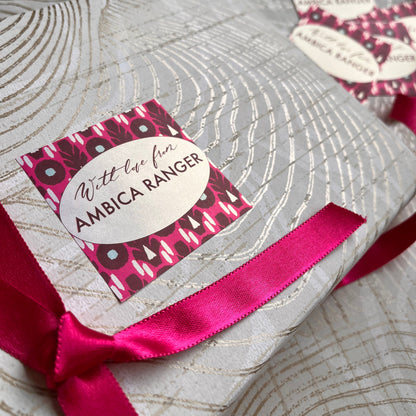 A close-up of a wrapped gift featuring a silver and gray patterned paper, tied with a bright pink satin ribbon and decorated with a matching pink and black patterned sticker that reads 'With the love AMBICA RANGER'. A second matching sticker and gold scissors are visible in the background.