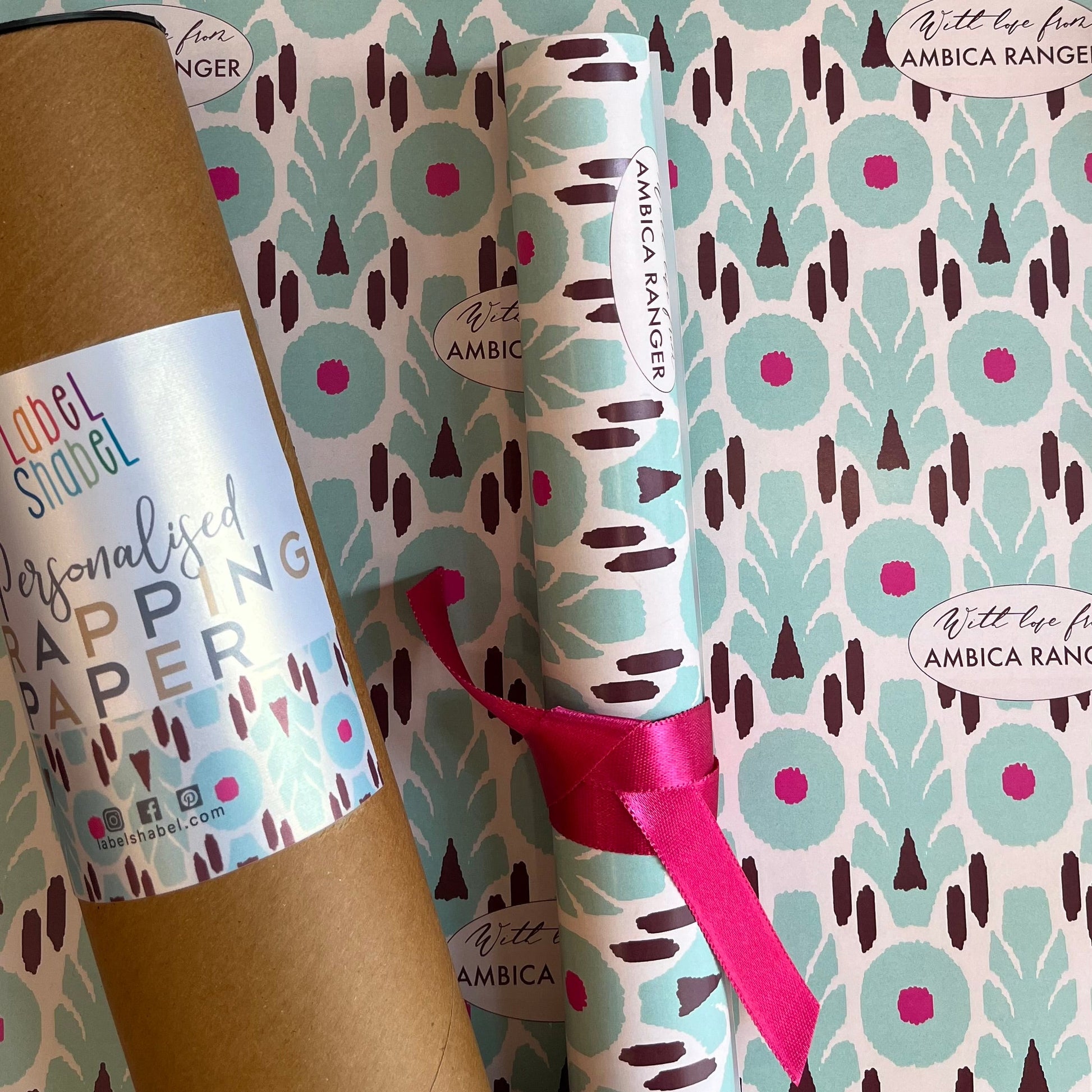 Personalized wrapping paper set by Amrica Ranger, featuring a kraft paper tube and a rolled sheet of the patterned paper tied with a pink ribbon, displayed against the patterned paper background.