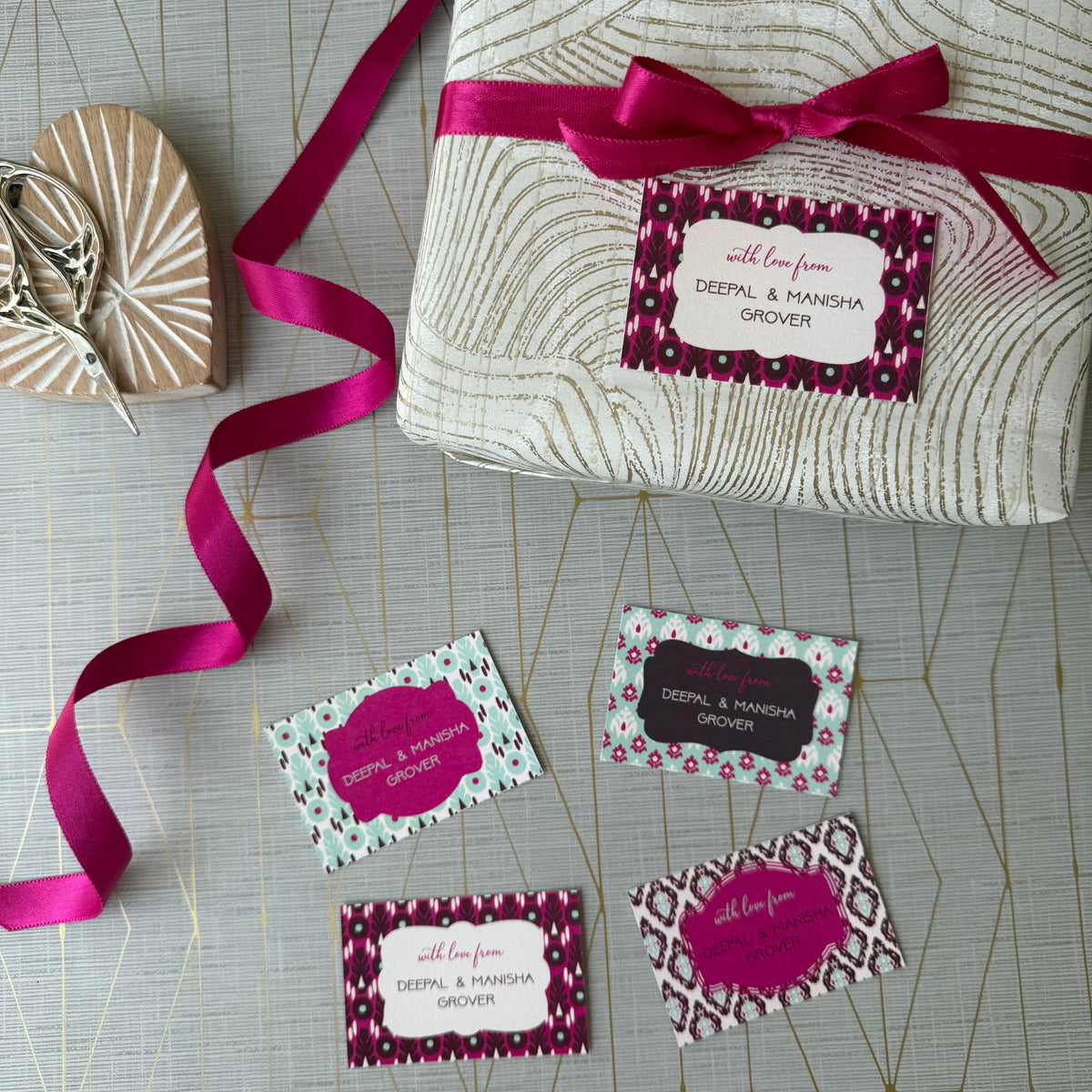 A wrapped gift box tied with a bright pink ribbon and a matching gift tag, arranged on a light gray patterned surface with several sample gift tags and a small wooden heart-shaped box nearby.
