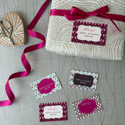 A wrapped gift box tied with a bright pink ribbon and a matching gift tag, arranged on a light gray patterned surface with several sample gift tags and a small wooden heart-shaped box nearby.