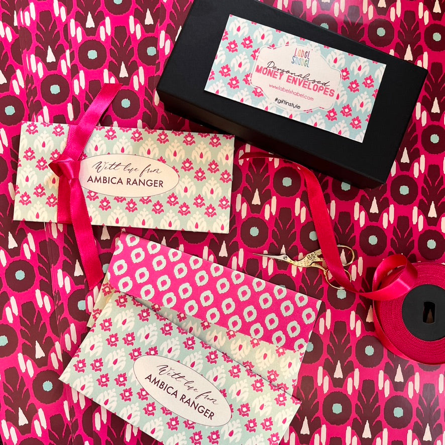 Money Envelopes gift set featuring three patterned envelopes with pink ribbons, a black presentation box, and a roll of pink ribbon on a matching patterned background.