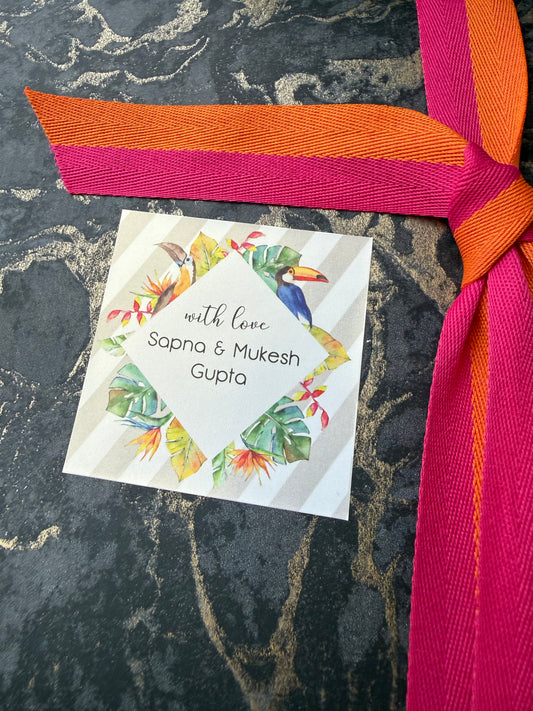A square white gift tag with tropical bird and leaf illustrations, tied with a bright pink and orange ribbon, placed on a dark marbled surface. The tag reads 'with love, Sapna & Mukesh Gupta'.