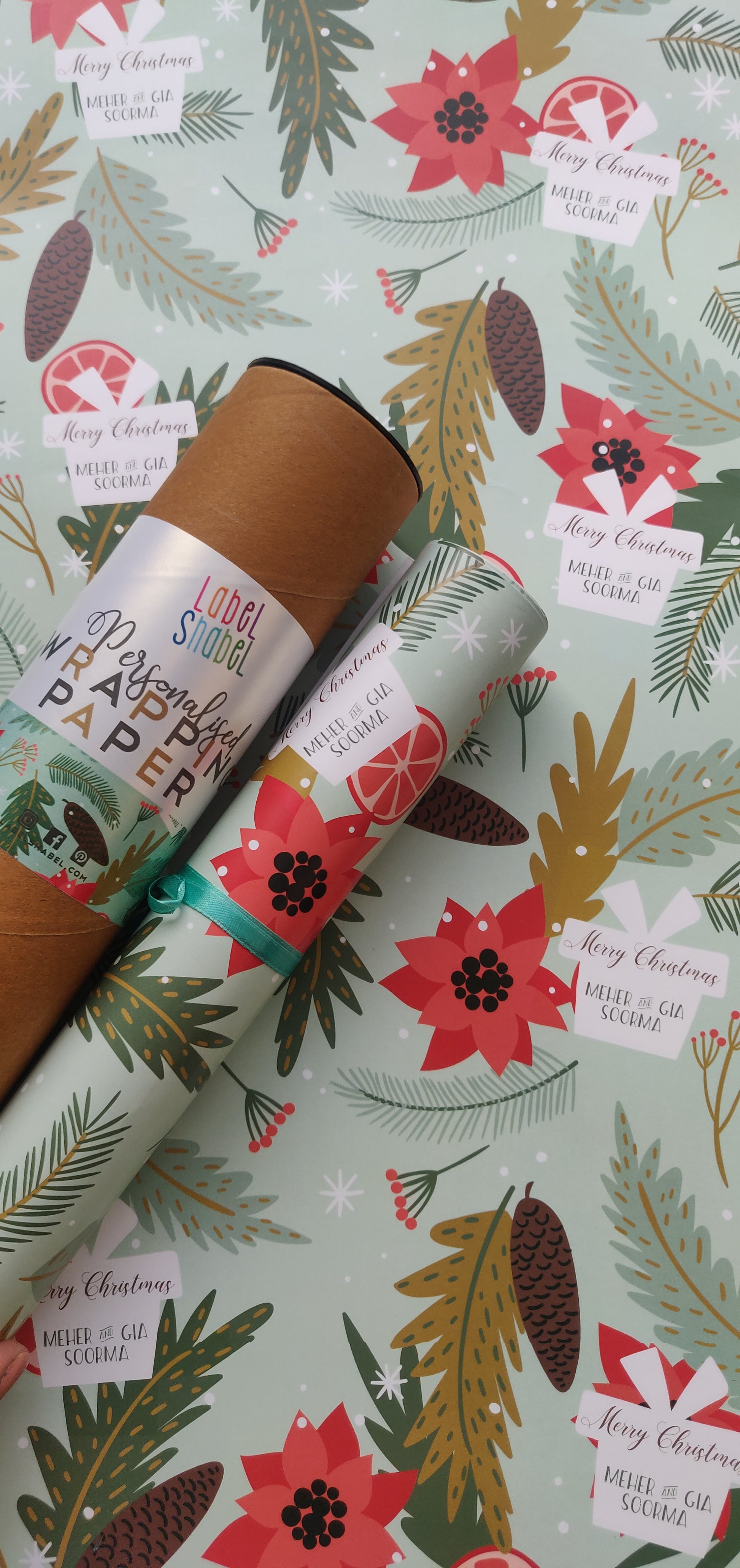 A roll of personalized wrapping paper with Christmas-themed designs including trees, berries, and snowflakes, placed next to a label that reads 'Merry Christmas'.