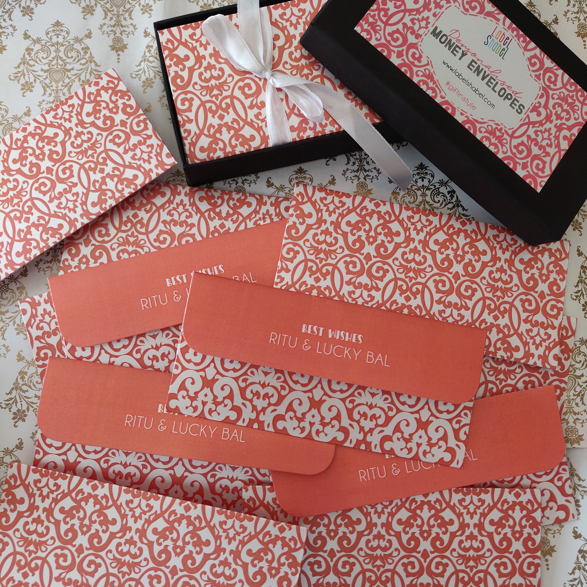 A flat-lay of money envelope gift set, featuring patterned envelopes, a black gift box with a white ribbon, and an open black envelope on a patterned fabric background.
