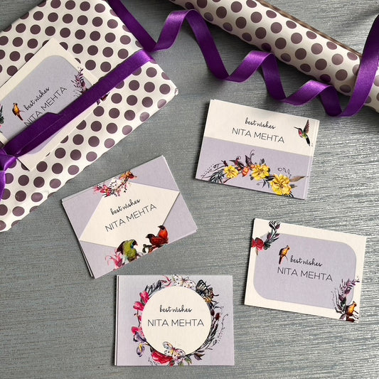 A flat lay of matching gift packaging and cards on a gray textured surface. The set includes a gift box wrapped in white paper with brown polka dots and a purple ribbon, a rolled matching paper tube, and several floral greeting cards with the text 'best wishes NITA MEHTA'.