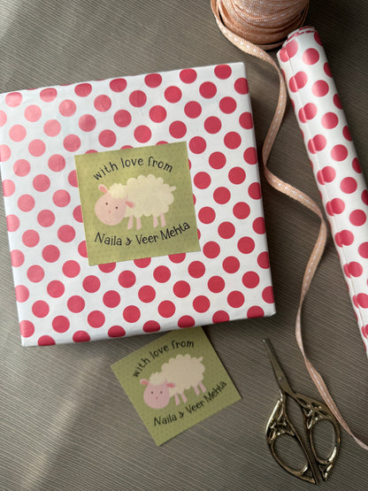 Personalized gift wrap featuring a pink and white polka dot pattern, wrapped around a square gift box with two custom tags. A matching roll of wrapping paper, a spool of ribbon, and a pair of scissors are arranged on a gray fabric background.