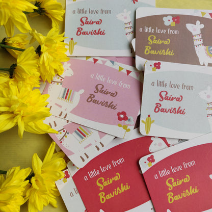 A styled flat-lay of printed cards featuring the phrase 'a little love from Sairo Bavishi' in different colorways, arranged alongside bright yellow flowers on a yellow surface.