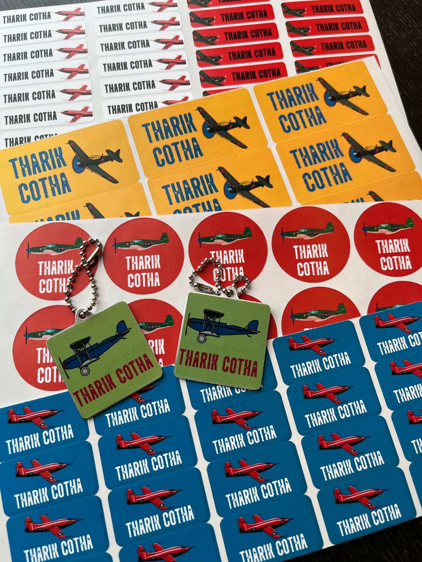 A sheet of colorful sticker sheets featuring a name with red, yellow, blue, and green airplane illustrations. Two small square keychains with matching stickers are displayed in front on a dark surface.