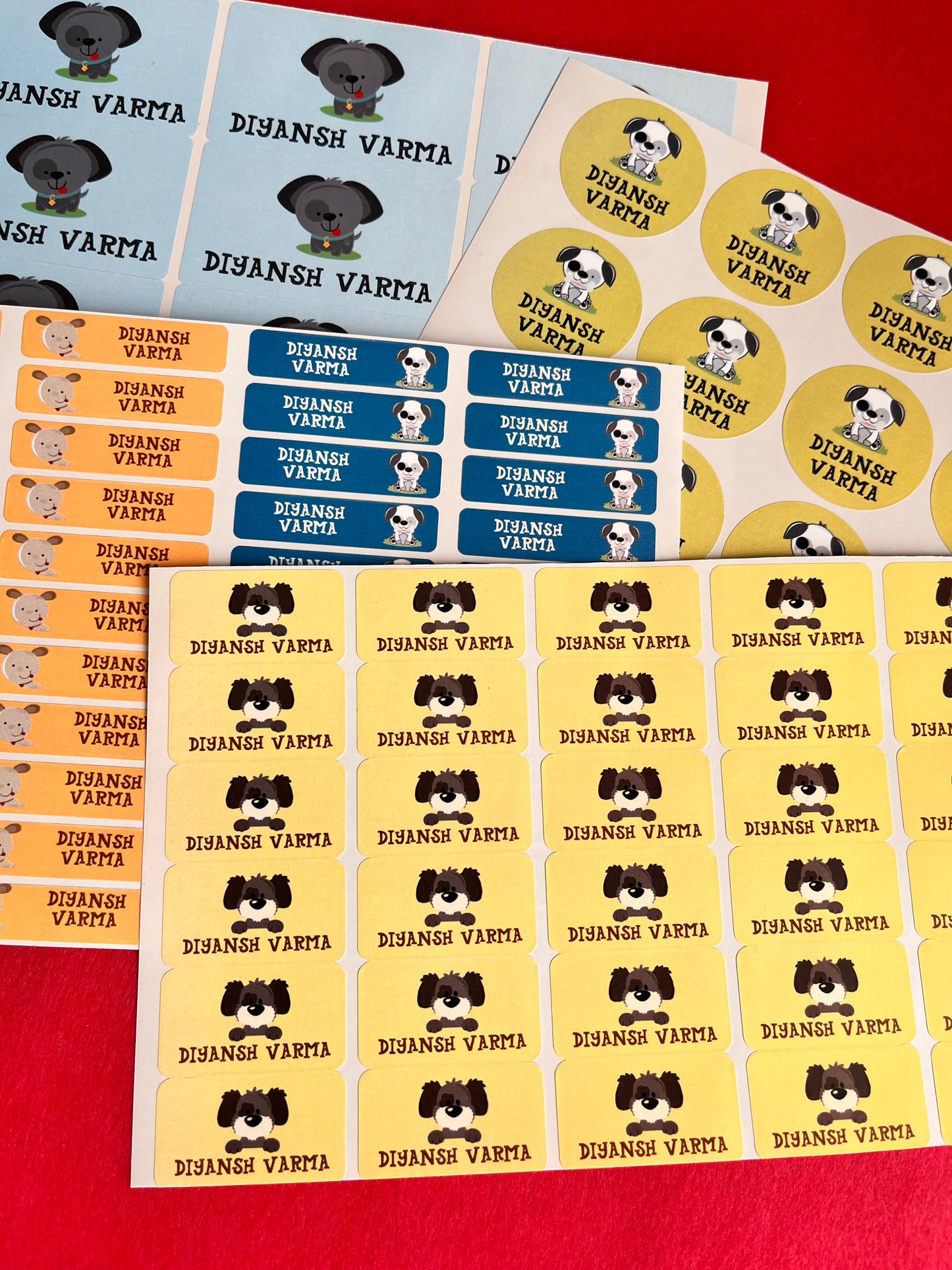 A set of colorful printed stickers featuring a name  and a cartoon dog illustration, arranged on a red surface. The stickers come in yellow, blue, orange, and white sheets.