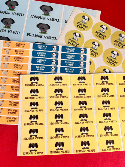 A set of colorful printed stickers featuring a name  and a cartoon dog illustration, arranged on a red surface. The stickers come in yellow, blue, orange, and white sheets.