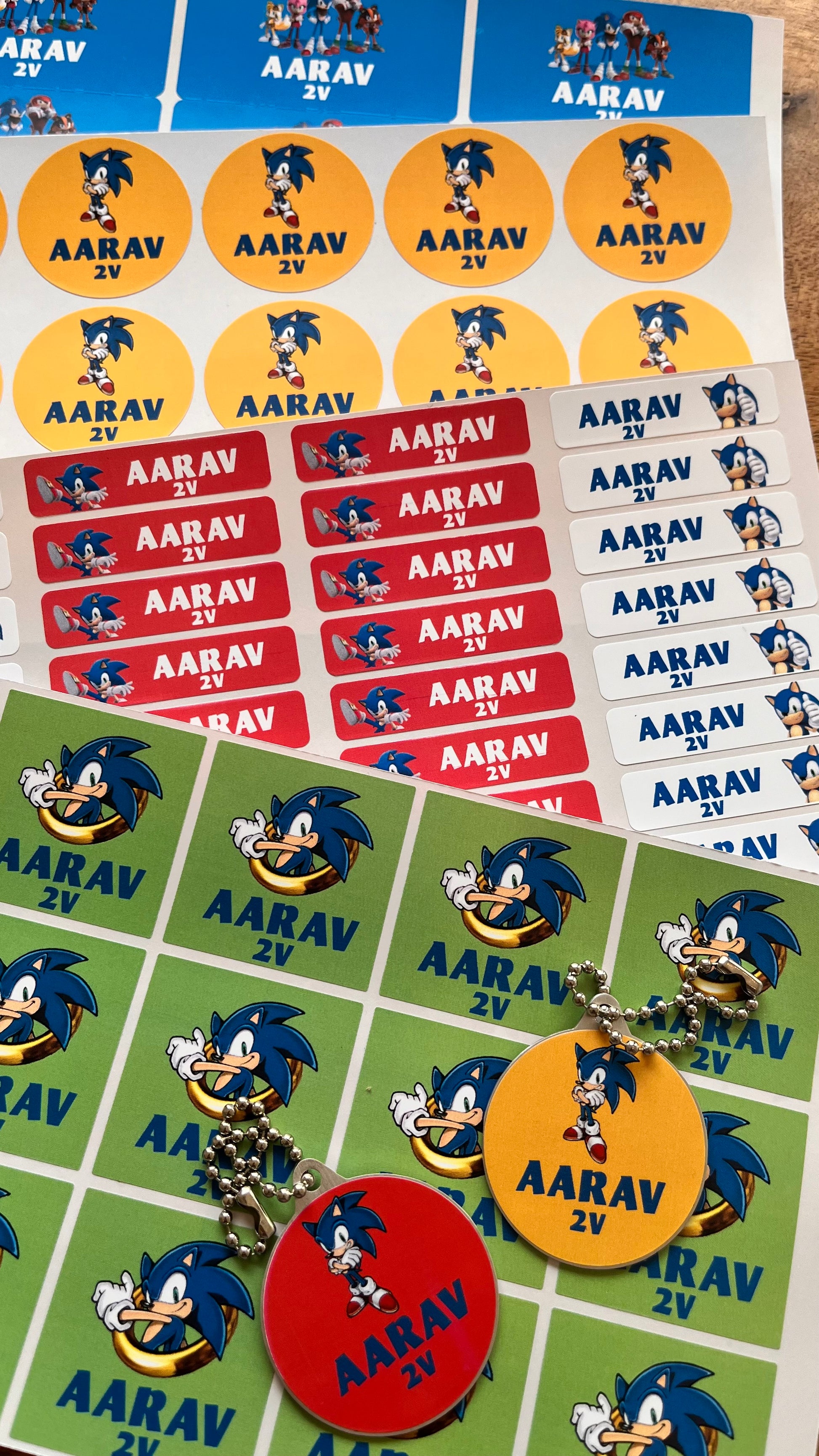 A collection of custom printed sticker sheets and keychains featuring Sonic the Hedgehog and the name 'AARAV 2V' on various colored backgrounds.
