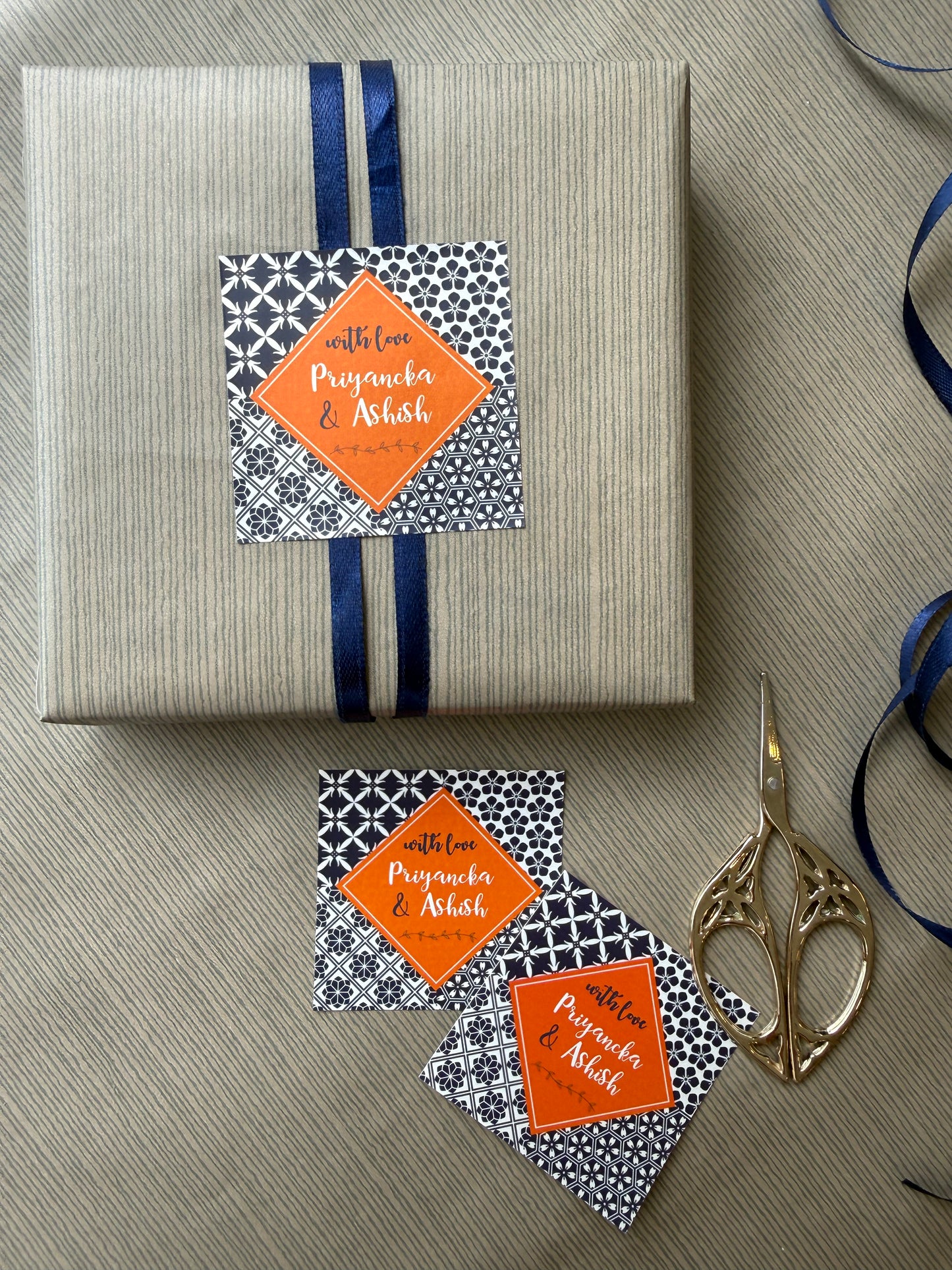 A wrapped gift box and matching paper tags with orange diamond labels, arranged on a textured beige surface with blue ribbon and gold scissors.