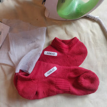 Red socks with 'ABhay' label on a beige surface next to a green and white clothes iron