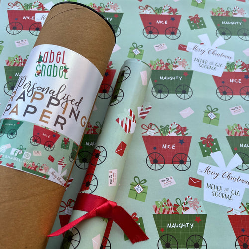 A roll of personalized wrapping paper with Christmas-themed patterns, including the words 'Naughty or Nice?' and 'Merry Christmas'. The paper is wrapped around a cardboard tube with a red ribbon and a label that reads 'Personalised Wrapping Paper'.