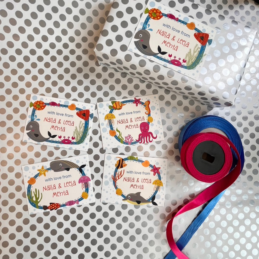Personalized underwater-themed gift tags and a roll of ribbon on a polka-dotted background. The tags feature colorful sea creatures like fish, octopus, shark, and whale, with the text 'with love from: Nabil & Leila Mona'.