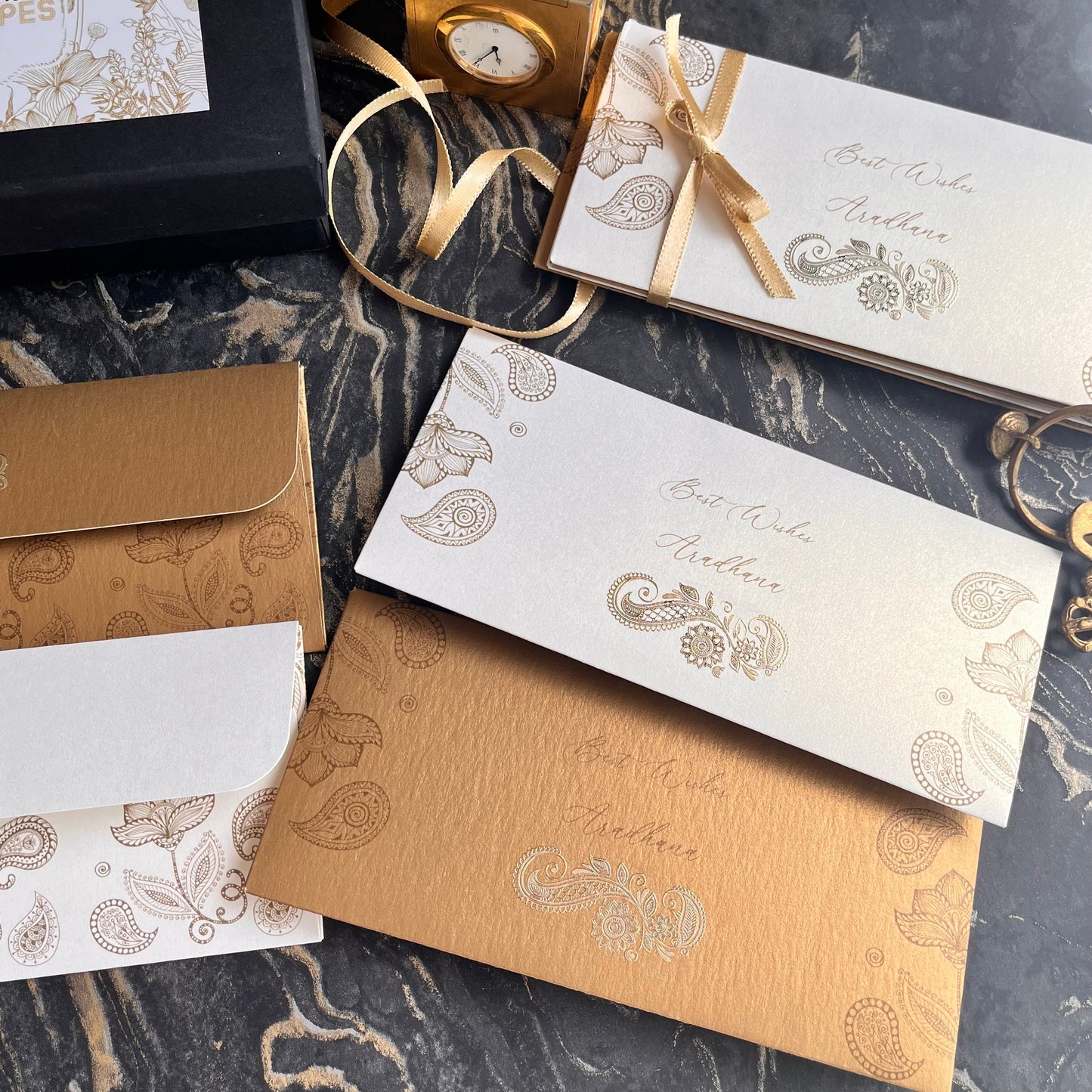 A collection of elegant gift envelopes with paisley patterns, gold accents, and a mix of silver and gold colors, displayed on a marble surface.