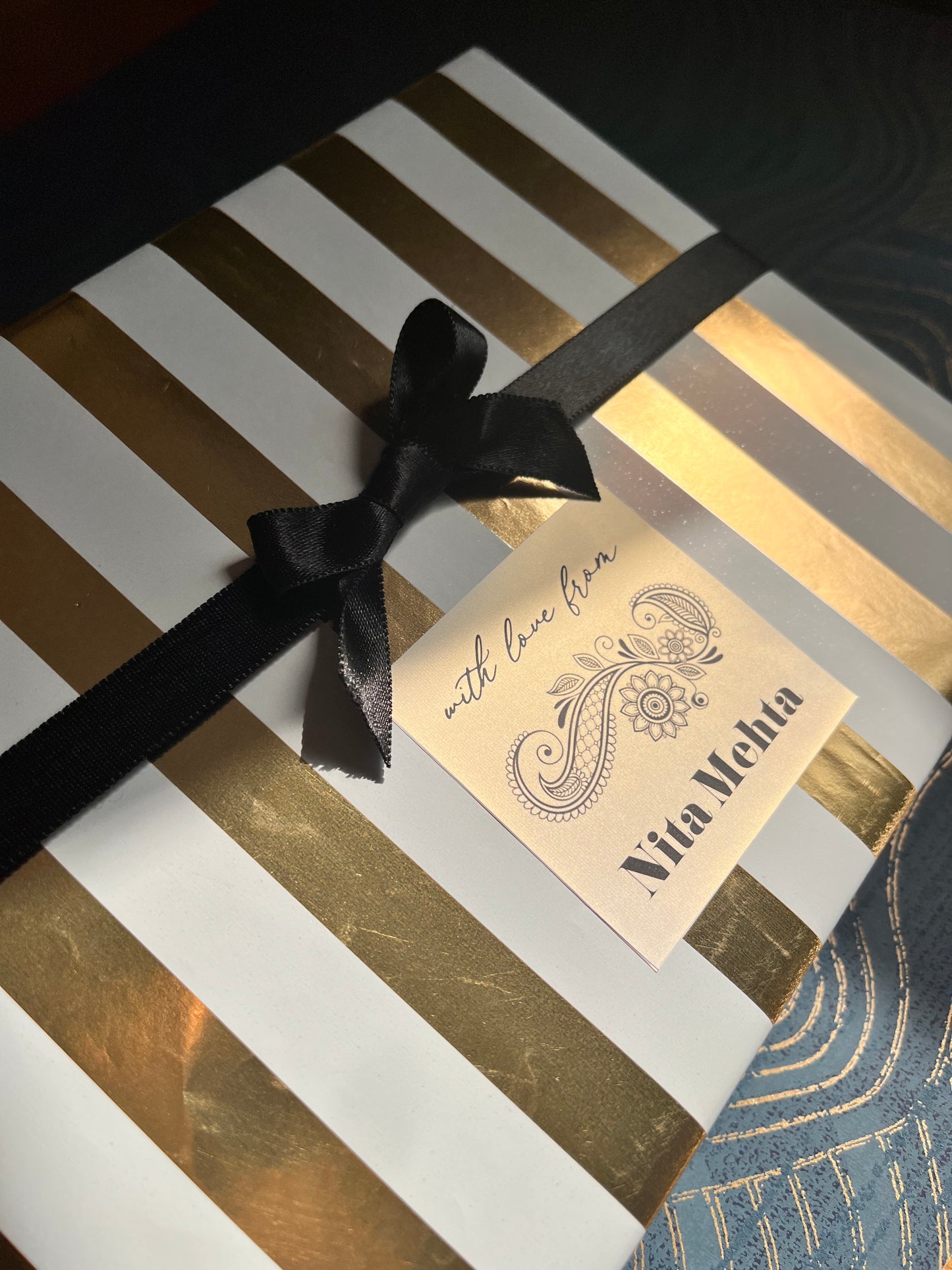 A close-up of a gift box wrapped in white paper with metallic gold stripes, tied with a black ribbon bow, and featuring a cream gift tag