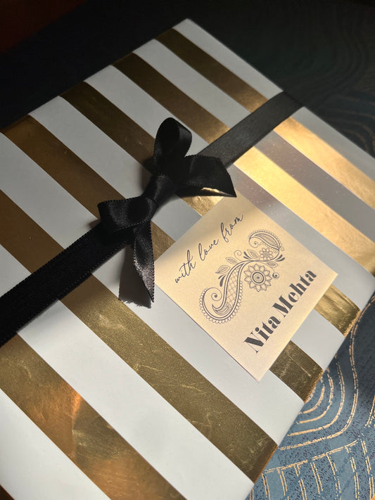A close-up of a gift box wrapped in white paper with metallic gold stripes, tied with a black ribbon bow, and featuring a cream gift tag
