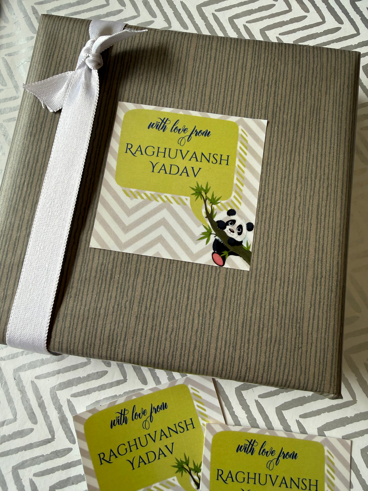 A wrapped gift box with a white ribbon bow, featuring a printed personalized card that reads 'with love from RAGHUVANSH YADAV' with a panda illustration. Two matching cards are placed in front on a gray and white patterned fabric background.