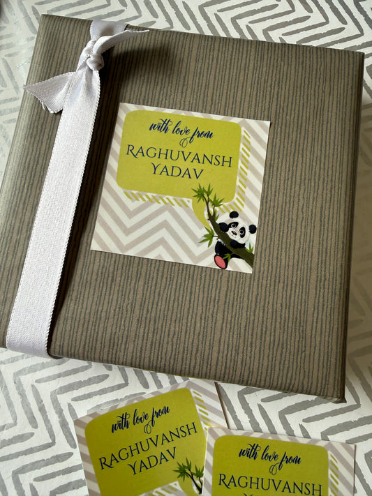 A wrapped gift box with a white ribbon bow, featuring a printed personalized card that reads 'with love from RAGHUVANSH YADAV' with a panda illustration. Two matching cards are placed in front on a gray and white patterned fabric background.