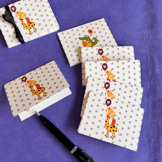 A set of children's greeting cards and matching envelopes with a cartoon giraffe holding a purple heart-shaped balloon, arranged on a textured purple fabric background next to a black pen.