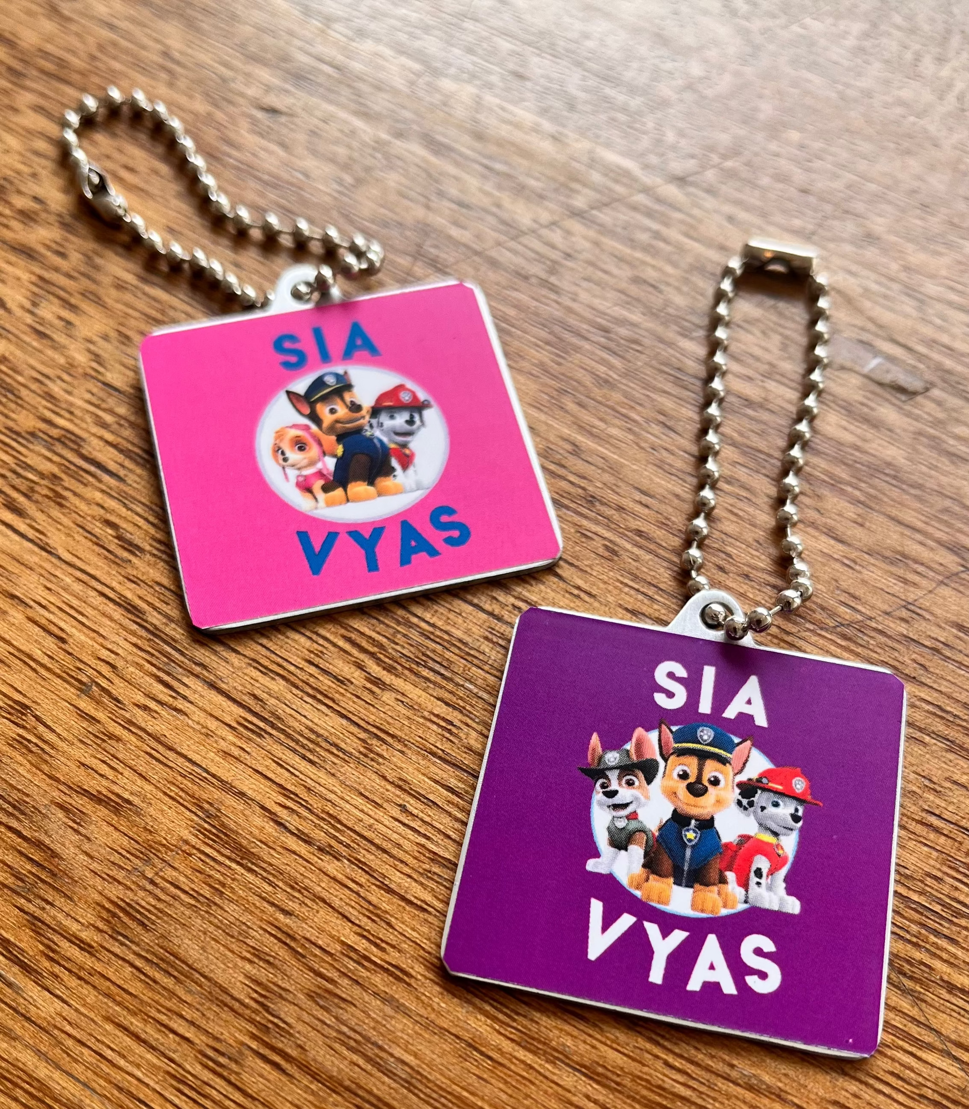 Two custom square metal keychains with Paw Patrol character graphics and the text 'SIA VYAS' in pink and purple, resting on a wooden table.