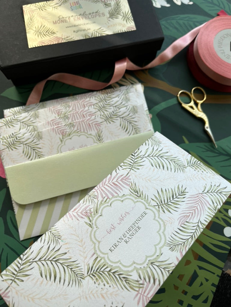 A styled flat-lay of  MOney envelopes Envelopes, showing a black branded box, a pale green envelope, a patterned envelope, pink ribbon, and gold bird scissors on a tropical green background.