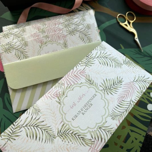 A styled flat-lay of  MOney envelopes Envelopes, showing a black branded box, a pale green envelope, a patterned envelope, pink ribbon, and gold bird scissors on a tropical green background.