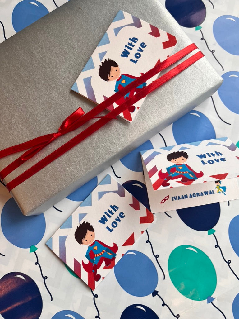 A wrapped gift box and two matching gift tags with a cartoon superhero illustration and the words 'with Love,' tied with red ribbons, placed on a white gift bag printed with blue and teal balloons.