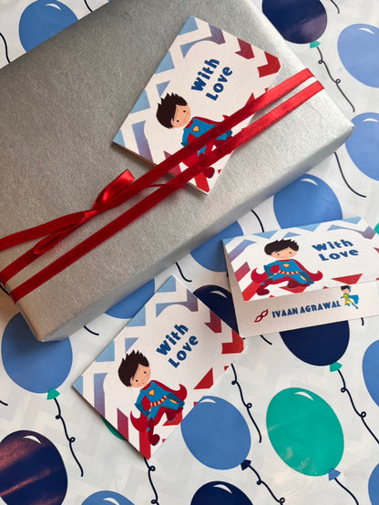 A wrapped gift box and two matching gift tags with a cartoon superhero illustration and the words 'with Love,' tied with red ribbons, placed on a white gift bag printed with blue and teal balloons.