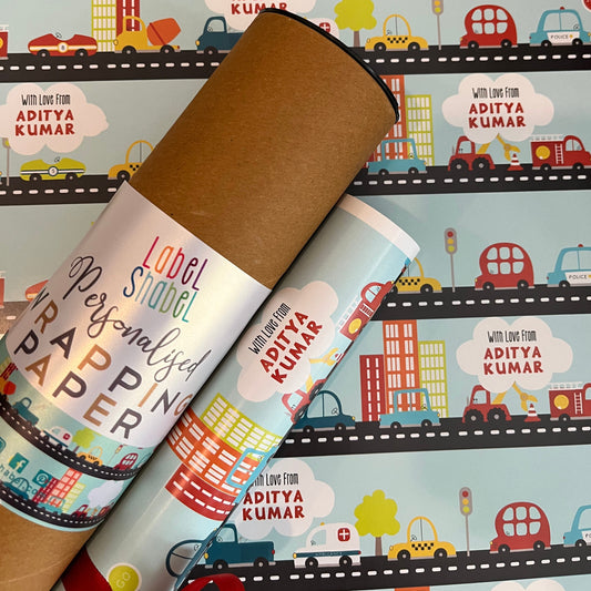 Personalized wrapping paper rolls displayed diagonally over a sheet of the same colorful city-themed wrapping paper. The rolls are wrapped in brown kraft paper with white labels that read 