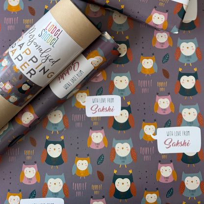 Owl-themed wrapping paper and gift wrap set with a repeating pattern of cute owls on a dark gray background, shown wrapped around gift boxes and a roll with a red ribbon.